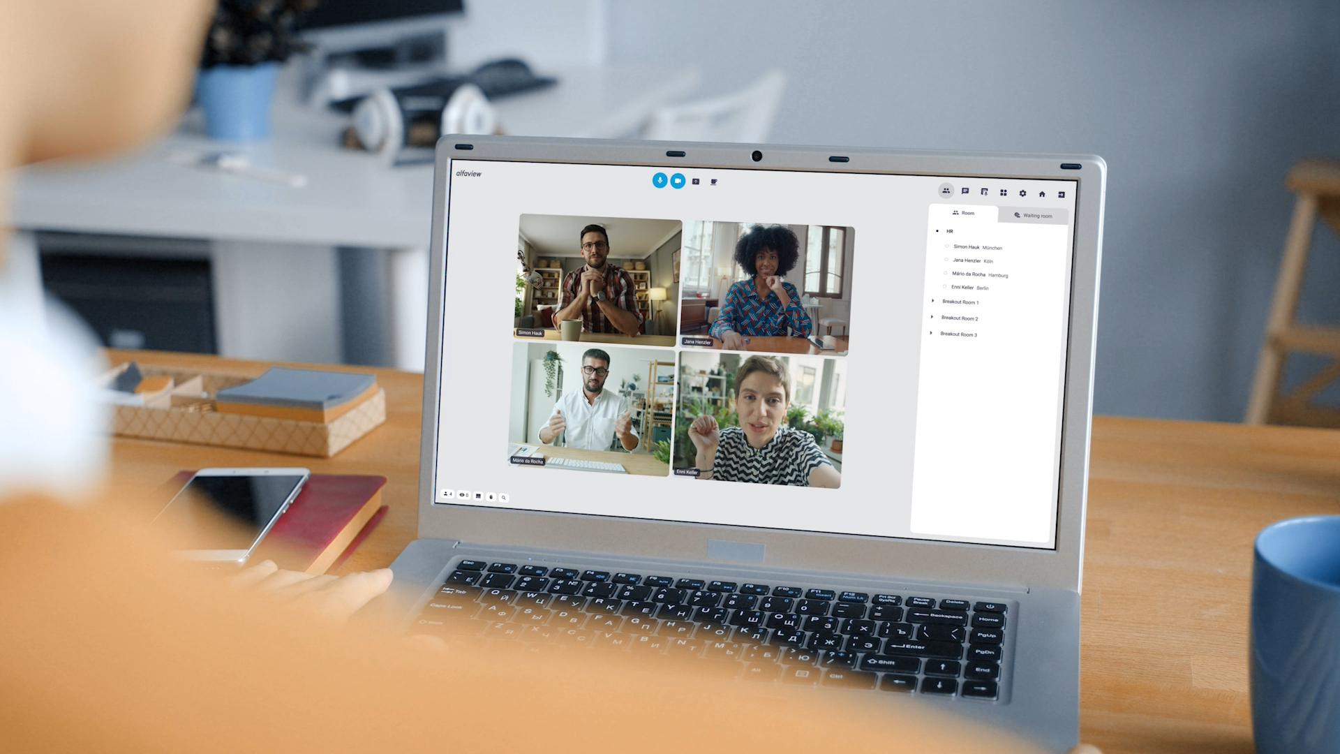 Film showing several people working together in alfaview. The people are in a video conference. The backgrounds show that some are working from home and others are in the office. The room structure in alfaview is shown with different rooms, each named after a different department. A number next to the room name shows how many people are currently in the room. Clicking on the room name takes you to another room. After exchanging a private short message, two people meet in a breakout room. They share the image from a second camera and work with a virtual whiteboard. This is followed by a video conference with video images of over one hundred participants. Small windows appear on the screen with live transcription and live translation. Finally, a woman from the home office waves to her colleagues, with whom she is connected via alfaview on her laptop.