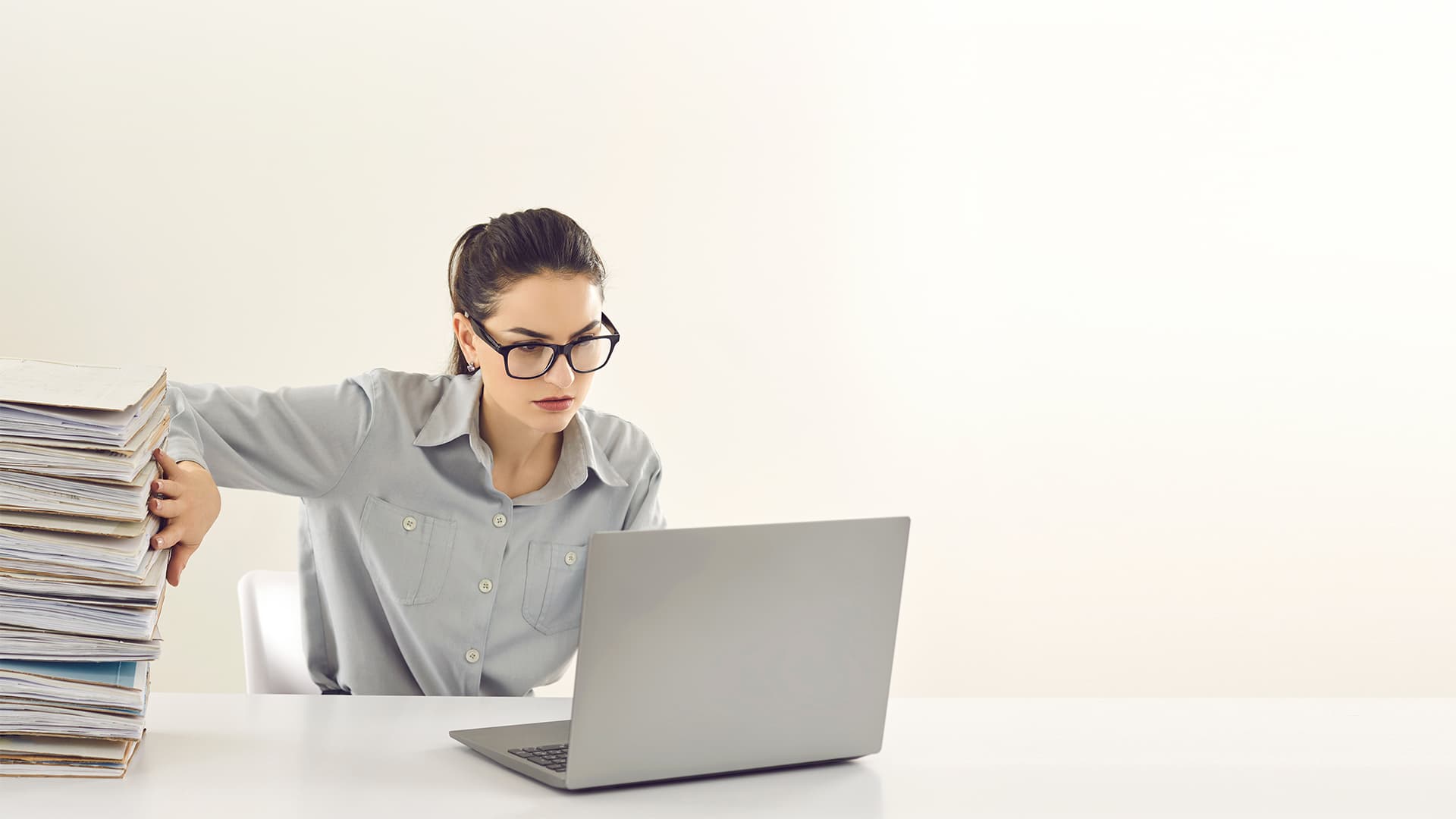 Woman sitting in front of laptop, pushing a pile of files off the desk with her right hand.