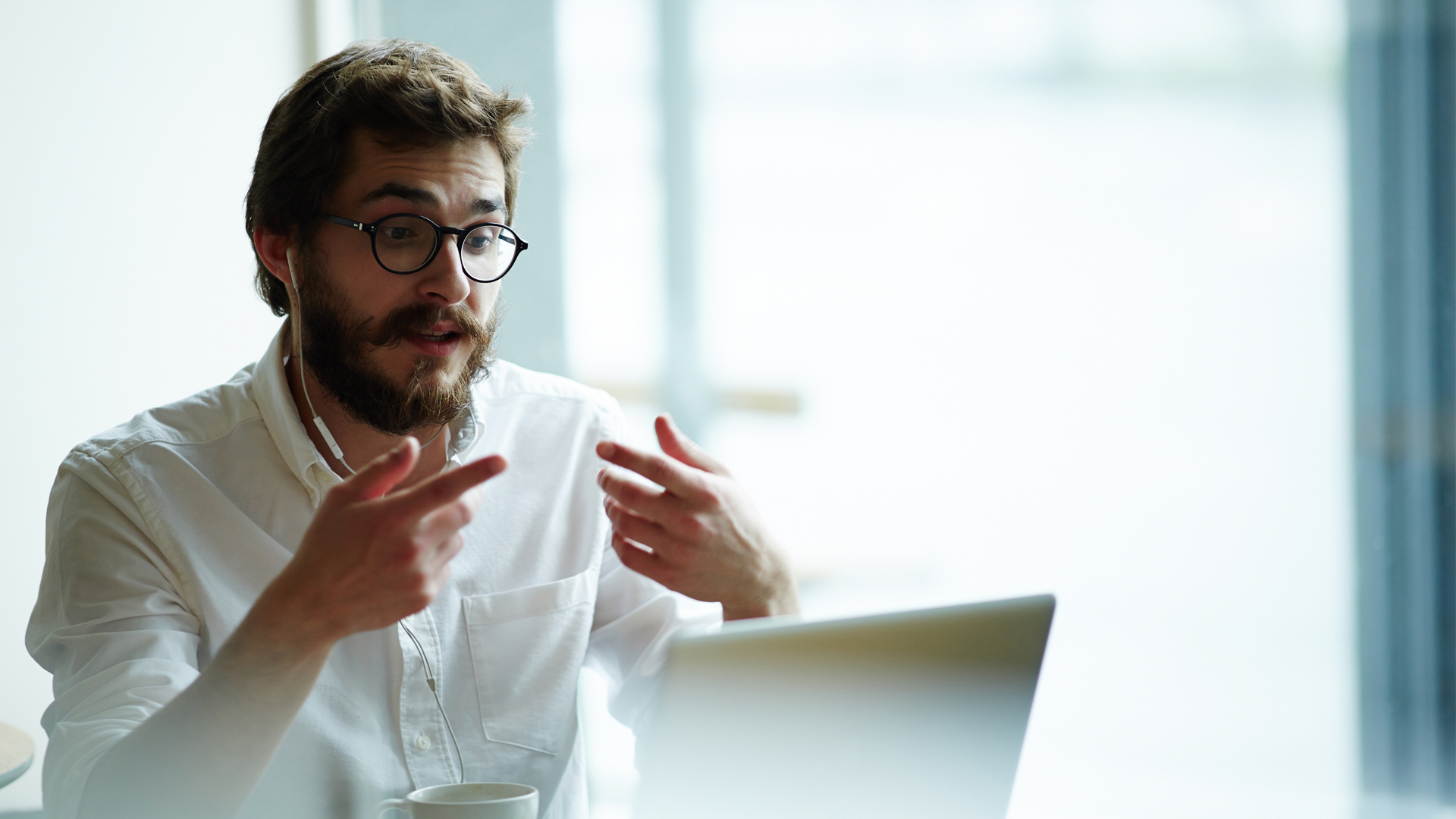 man sits in front of a laptop wearing earplugs and gesticulating with his hands.